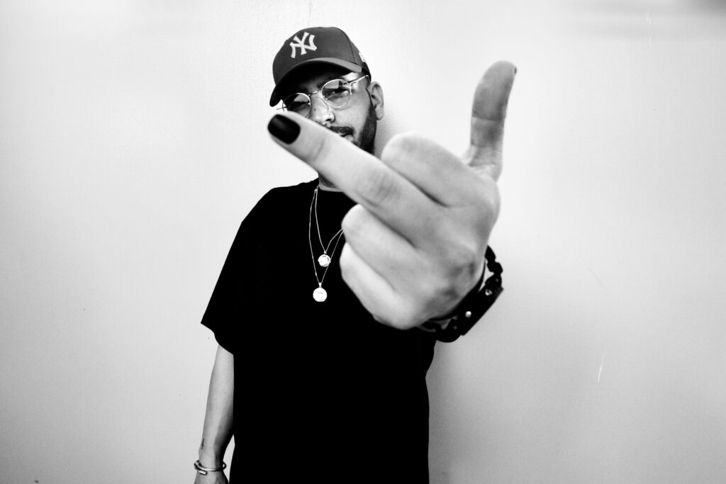 Fotografia en blanco y negro de un chico joven con gafas modernas enseñando el dedo corazón en primer plano. El chico lleva gorra y las uñas pintadas.